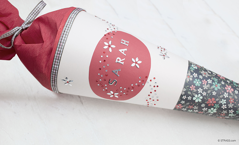 Weiße Schultüte mit Blumenpapier, rotem Namensfeld und kleinen Kristallen.