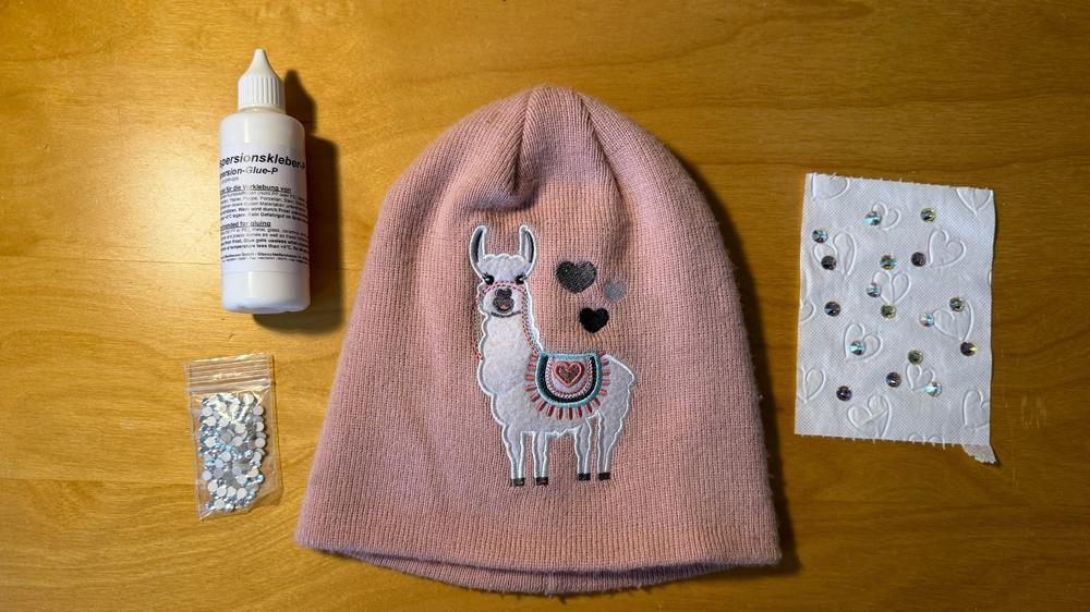 Rosa Mütze mit Lama-Motiv, Strasssteine, Strasskleber und Pick-up-Stift als Material für die Bastelanleitung.